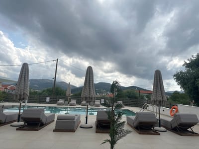 The image captures an outdoor poolside area, likely at a resort or hotel, under a heavily overcast sky. In the foreground, a light-colored tiled patio extends across the scene, furnished with multiple modern gray sun loungers, some featuring adjustable backs and wooden frames. Four tall, closed sun umbrellas with a distinctive woven or macrame-like texture are evenly spaced along the pool's edge. A small, sparsely branched evergreen plant in a pot stands centrally in the foreground.
The mid-ground features a rectangular swimming pool with clear blue water. Metal handrails are visible on the right side of the pool, and an orange and white lifebuoy hangs on a white wall nearby. Behind the pool, a dark wooden slatted fence acts as a boundary. Beyond this fence, several gray beanbag-style chairs and small tables are visible.
The background reveals a natural landscape with rolling hills and mountains, covered in scattered greenery, extending under the cloud cover. Small residential buildings are nestled among the distant terrain. A wooden utility pole with overhead wires is present on the left side of the scene. On the far right, a section of a large green tree is visible.
The sky is dominated by dense, dark gray clouds, though a small area on the far left shows a lighter sky with white cumulus clouds, suggesting a partial break in the overcast conditions. The overall impression is a daytime scene marked by gloomy or subdued weather. A small white sign with pictograms, likely indicating pool rules, is affixed to the dark wooden fence to the left of the pool's center. No people are visible, and no specific activity is taking place. The setting evokes a resort environment in a hilly region, consistent with Limenaria, Greece.
