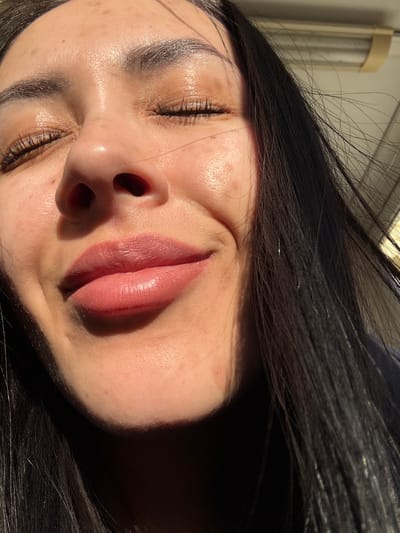 A close-up, indoor photograph, taken at an oblique angle, features a woman with closed eyes. Her face is predominantly bathed in sunlight, creating strong contrasts of light and shadow. She has dark hair, full lips with a rose-nude lipstick or gloss, and well-defined eyebrows. The background is indistinct, but hints of a window or light fixture are visible in the upper left corner.
The location in the city of Molodezhnyi, Russia, is not discernible from the image.