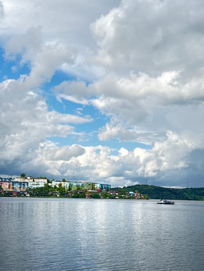 The image depicts a waterfront scene with a town lining the shore. The sky is filled with dramatic, voluminous clouds, some white and fluffy, others dark and heavy, interspersed with patches of bright blue sky. A body of calm water stretches out to the right of the frame, reflecting the light and appearing textured.

Along the shore, a row of buildings with colorful facades creates a vibrant strip of urban development. The buildings are of varying heights and styles, with visible rooftops and windows. Green trees are interspersed among the buildings, suggesting a lush environment. In the distance, a larger, dark green, hilly landscape can be seen. A boat with several people on board is visible on the water, heading away from the shore.

The scene suggests a daytime setting, likely late morning or early afternoon, given the brightness of the sky. The weather appears mixed, with a mix of sun and clouds. The location appears to be a coastal or lakeside town. There is no visible text in the image.