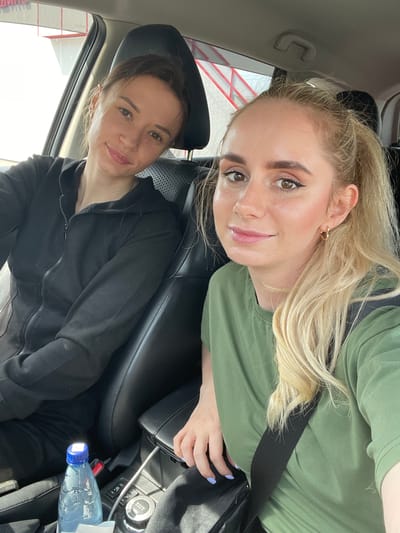 Two women are seen inside a car, captured in a selfie-style photograph during what appears to be daytime. The image is oriented vertically with the subjects looking towards the camera.
The woman in the foreground has blonde hair, styled with some strands pulled back, and is wearing a green short-sleeved top with a black seatbelt across her chest. She has prominent dark eyebrows, noticeable eyeliner, and subtle makeup, including light pink lipstick. She wears a small gold hoop earring in her visible ear and has light blue nail polish on her left hand, which is resting near her lap. She is smiling gently.
Behind her, in the passenger seat, is another woman with dark hair, which appears to be tied back. She is wearing a black zippered hoodie and also has a subtle smile, looking towards the camera.
The car interior features black leather seats and headrests. On the center console, between the two women, a part of the dashboard with controls is visible, including a knob with "4x4 AUTO" text. A silver cable is plugged in nearby. A plastic water bottle with a blue cap is resting on the console or floor area.
Outside the car window, through the bright daylight, some red and white structures are visible, possibly part of a building or an industrial area. Faint red Cyrillic text can be seen on a white background in the lower left window area, and also partially through the rear window, hinting at commercial or public signage. The overall mood conveyed by the women's expressions is relaxed and pleasant. The scene takes place in Kiparisovo-2, Russia.