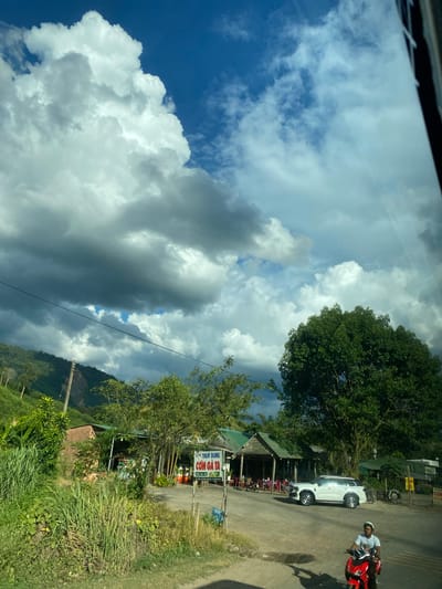The image shows a rural scene in Thôn 18, Vietnam, under a partly cloudy sky. In the foreground, a person on a red motorcycle wears a white helmet and a white t-shirt. To the left, lush green vegetation, including tall grass and bushes, grows abundantly. In the middle ground, a dirt road leads past several open-air buildings with green roofs, resembling small shops or restaurants. A white SUV is parked on the road. A sign with Vietnamese text, including "CƠM GÀ TA", is visible near the buildings. In the background, a densely wooded hillside rises, with some buildings and power lines visible on the slopes. The sky is a mix of bright blue and dramatic dark clouds, suggesting a dynamic weather system. The overall impression is of a roadside stop in a verdant, mountainous region.