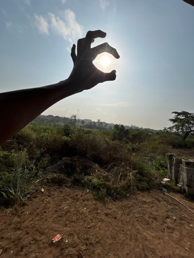 The image displays an outdoor scene, reportedly in Debasco, Nigeria, under a clear, bright sky with scattered thin clouds. A human left arm and hand are silhouetted in the upper left quadrant, with the hand cupped around the intensely bright, low-angle sun, which emits visible rays. The foreground consists of reddish-brown, dry, disturbed earth, featuring scattered small debris, including a red wrapper, and patches of dry grass. The midground is dominated by dense, unkempt vegetation, comprising various green and dry shrubs and tall grasses. In the background, a hazy landscape shows the faint outlines of structures, likely buildings, and a few tall trees, including a distinct palm tree. On the far right of the foreground, a stack of grey concrete blocks is visible alongside a light-colored pipe or hose lying on the ground.