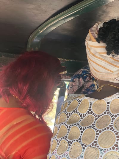 This image depicts the interior of a vehicle in Awjaw, Nigeria, showing three individuals from a rear-side perspective. In the foreground on the left, a person with short, wavy, bright red hair wears a light orange top with subtle horizontal stripes and a thin gold-colored necklace. Adjacent to them on the right is another person wearing a white garment with a prominent gold-and-silver beaded circular lace pattern, a gold-colored necklace, and a cream-colored headwrap with orange and white stripes covering dark, curly hair. Partially visible between these two individuals in the background is a third person wearing a blue patterned garment. The vehicle's dark ceiling and a green metal support bar are visible at the top of the frame. The scene is illuminated by natural light, suggesting daytime.