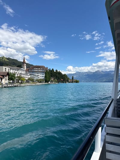 A view from a boat on a bright, sunny day shows the clear, turquoise waters of Lake Thun in Hünibach, Switzerland. In the distance, a charming Swiss town lines the shore, featuring a prominent church tower with a red steeple and clock face. The town is characterized by its white, multi-story buildings, many with balconies and intricate architectural details, nestled against a backdrop of lush green hills and distant, majestic mountains. The boat's railing and seating are visible in the foreground, indicating the perspective of someone on board. The sky is a vibrant blue, dotted with fluffy white clouds, and the water exhibits gentle ripples and waves.