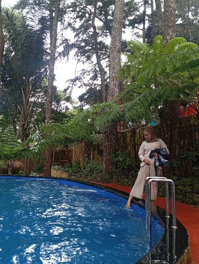 A female individual is observed standing on the reddish-orange paved deck of an outdoor swimming pool located in Kuwukan, Indonesia. The pool features a blue interior lining and black coping, with visible water surface agitation. The subject is attired in a light-colored, possibly beige, striped long-sleeved top, matching wide-legged pants, and a light-brown hijab. She holds a folded, dark-colored textile object in her left arm. Her right foot is partially submerged in the pool water, adjacent to stainless steel pool ladder handrails. The environment is characterized by dense, tall green trees, including coniferous and broadleaf types, and abundant fern foliage, forming a naturalistic backdrop. A rustic-style fence or wall is visible behind the pool area, constructed from vertical wooden or bamboo slats, with rocks and additional vegetation at its base. The lighting indicates an overcast sky or diffused light conditions.