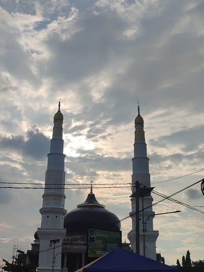 The image shows the facade of a mosque in Keudematangglumpangdua, Indonesia. The mosque has two tall minarets with golden domes and a central dome. The sky is cloudy with the sun peeking through the clouds. There are power lines in the foreground. The mosque appears to be under construction, as evidenced by scaffolding on the left side of the building. A blue tarp is visible in the lower part of the frame. A sign with text in Indonesian is also present.