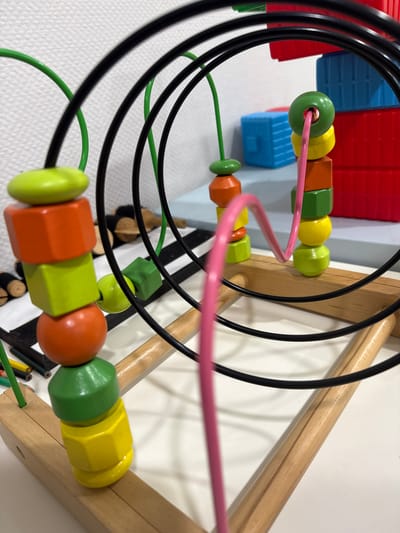 The image shows a close-up of a wooden bead toy with colorful beads on wire loops. The beads are stacked in various shapes and colors, including green, orange, yellow, and red. A pink wire loop goes through some of the beads. In the background, there are stacked red and blue plastic blocks. The scene is set against a white, textured wall. There is no indication of the city of Sultanbeyli, Turkey, or any people in the image. The lighting suggests an indoor environment.