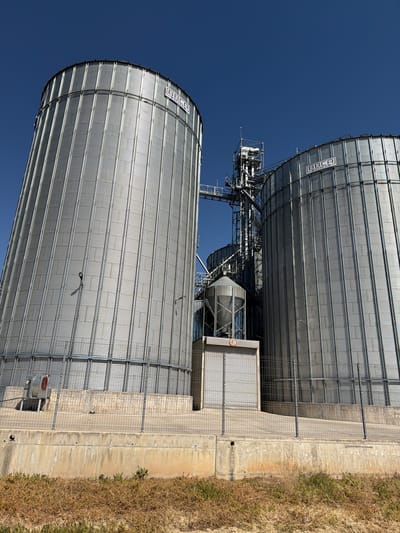 Two large metal grain silos stand near Umarevtsi, Bulgaria. They are cylindrical, corrugated, and silver-colored, with the branding "BBCA" visible on the top of each. A network of metal walkways and platforms connects the silos. A small gray building is adjacent to one silo, enclosed by a chain-link fence. The ground consists of a concrete pad bordered by dry grass. The sky is clear and deep blue.