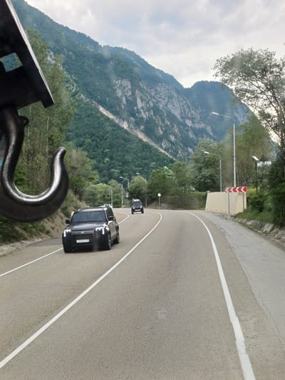 The image captures a winding mountain road in Monastyr, Russia, under a cloudy sky. The perspective is from a slightly elevated position, looking down at the road and the surrounding landscape.
The primary subjects are two black SUVs driving on the asphalt road, marked with white lines. The lead SUV is closer to the foreground and is more detailed, showing its license plate which reads "D555PM 123". The second SUV is further up the road. The road itself curves to the right, disappearing into the distance.
The setting is a mountainous region with lush green trees covering the steep slopes. The mountains are rocky and imposing, with some areas showing bare rock faces. The sky is overcast, suggesting a potentially cool or damp day. The presence of streetlights along the road indicates it is likely a developed route. In the foreground, a large, dark metal hook is visible, suggesting the image might have been taken from a vehicle equipped with some sort of lifting or towing apparatus.
The activity is simply vehicles traveling along a scenic mountain road. There are no people clearly visible in the image. The overall impression is one of a journey through a natural, somewhat remote landscape. No specific emotions are conveyed due to the absence of human subjects and the nature of the scene.