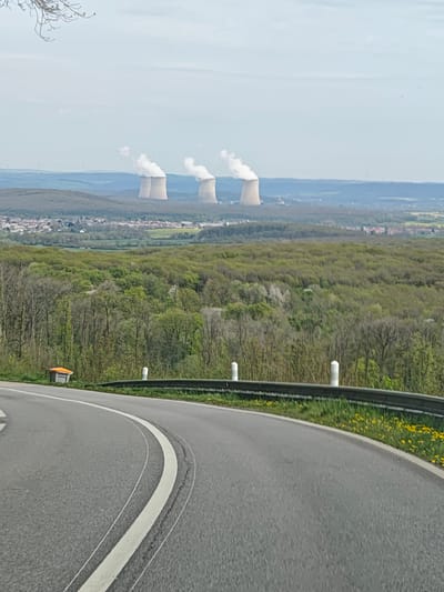 The image shows a view from a winding road overlooking the town of Thionville, France. In the distance, three nuclear power plant cooling towers are visible, emitting steam. A forest and fields fill the space between the road and the town. The road has a double white line marking the edge of the lane and a metal guardrail with white posts. Dandelions are growing along the roadside. A small orange and white container is visible next to the guardrail. The sky is overcast.