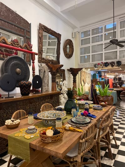 A long wooden table set for a meal with wicker chairs lining the sides, placed on a black and white checkered floor. Table settings include plates, glasses, and decorative elements. The backdrop features a wall adorned with mirrors, wooden sculptures, and a ceiling fan. Various items, including gongs and decorative masks, are scattered throughout the space.