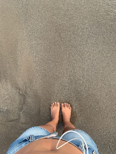 A first-person perspective shot shows bare feet standing on dark, wet sand. The individual is wearing blue denim shorts. White earbuds are visible, draped over the shorts. The scene appears to be a beach in North Kuta, Indonesia.