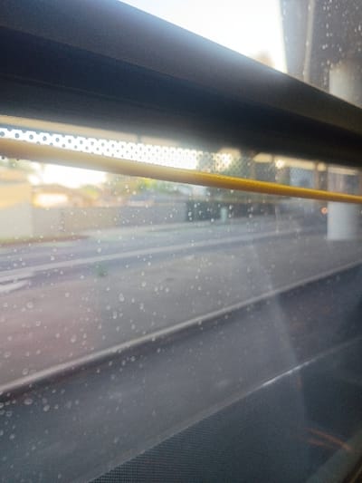 The image depicts an interior view from a public transport vehicle, specifically a light rail train, in Long Beach, United States. The perspective is directed outwards through a window, the glass of which is covered with numerous small water droplets. A dark, angular frame borders the top edge of the window. Below this frame, a horizontal, perforated light-colored panel is visible. A yellow, braided grab strap extends horizontally across the upper-middle section of the window, positioned internally. The exterior scene, appearing blurred due to motion or shallow depth of field, displays two parallel dark tracks, indicative of a light rail line, running horizontally across the mid-ground. A faint white line is discernible to the left of these tracks. Beyond the tracks, indistinct urban structures and sparse green foliage are present under a bright, potentially overcast, sky. The lower foreground of the image consists of dark, textured interior surfaces of the vehicle. No individuals are present or discernible within the frame.