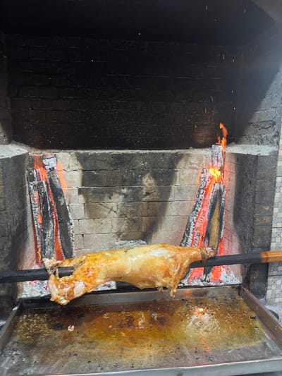 In a rustic setting, possibly a restaurant in Miatli, Russia, a whole lamb is being roasted over an open wood fire in a brick oven. The lamb, golden-brown and glistening, is skewered on a metal rod, positioned centrally in front of the flames. Burning logs sit on the oven floor, providing the heat source. To the right of the lamb is a greasy metal tray. The interior of the oven is lined with stone tiles, some stained black. The scene is dimly lit, with the fire casting an orange glow.