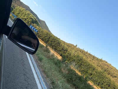 The image shows a rearview mirror reflection from a vehicle traveling on a road in Sant Feliu de Buixalleu, Spain. The reflection shows a person's arm with a bracelet and rings, and a partial view of the vehicle's interior. Outside the car, a tree-covered mountain slopes upwards under a clear blue sky. Roadside markers with blue and white chevron patterns are visible along the road, indicating a curve. The road surface is asphalt with white and blue lane markings. Grassy vegetation lines the side of the road, and the trees on the mountainside are dense and green. The lighting suggests it is daytime, likely late afternoon, with the sun casting shadows.