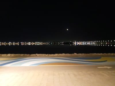 The image is a night-time, wide-angle shot of a waterfront promenade in Al Khobar, Saudi Arabia. The foreground features a paved walkway with a decorative, wave-like pattern in shades of blue, white, and yellow. A white arrow, indicating direction, is visible on the right side of the paved area. Beyond the walkway, a line of rocks separates it from a dark body of water. In the background, across the water, a city skyline is illuminated by numerous lights, reflecting on the water's surface. A crescent moon is visible in the dark night sky. The scene is devoid of any people or active events.