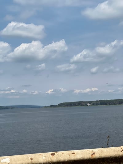 The image depicts a wide landscape view of the Pechinehi Reservoir, located near Staryi Saltiv, Ukraine. In the immediate foreground, a weathered, light-colored barrier, possibly concrete or metal, with significant rust and peeling paint, spans the lower portion of the frame. The midground is dominated by the expansive, grey-blue water of the reservoir, exhibiting subtle ripples across its surface. The distant right bank features a dense, dark green forest of coniferous trees. Within this treeline, a small, light-colored building with a dark, peaked roof is partially visible. The distant left bank appears as a low, indistinct landmass receding towards the horizon. The sky is light blue, occupied by numerous scattered white cumulus clouds. No people or active subjects are visible.