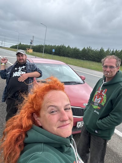 A man wearing a gray baseball cap, a checkered shirt over a black t-shirt with a "Linkin Park" graphic, and dark pants stands next to a red car. He is holding a cigarette in his right hand. To his right, another man in a green hoodie with a fox graphic and glasses, also smoking, stands next to the same car. In the foreground, a woman with bright red, curly hair, wearing a green hoodie, is partially visible. The scene appears to be on the side of a road, with trees and a cloudy sky in the background. A license plate on the car reads "P3....". The location is identified as Alnashi, Russia.