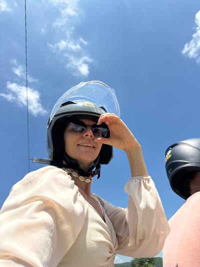 In a low-angle, close-up shot taken in Ham Thuan Bac, Vietnam, a woman wearing a black and grey motorcycle helmet with a clear visor smiles and shields her eyes with her right hand. She has dark sunglasses on and a necklace made of small white shells. She is wearing a light-colored, possibly cream-colored, blouse with puffed sleeves over a ribbed tank top. The sky is bright blue with scattered white clouds. A black helmet and a pink garment are visible in the background to the right, suggesting another person is present. The image appears to be a selfie taken while riding a motorcycle.