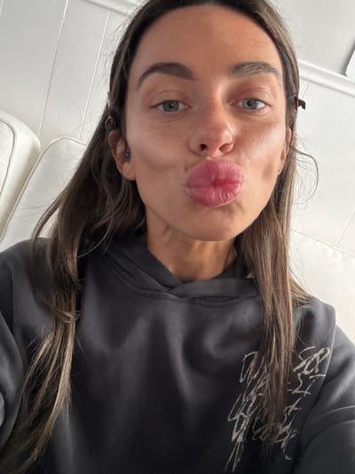 A young woman is captured in a close-up selfie, with her head tilted to the right and her body angled. She makes a playful puckered "kiss" face towards the camera. Her long, dark brown hair cascades over her shoulders and part of her dark grey or black hooded sweatshirt. She has striking blue-grey eyes, natural eyebrows, and a small silver hoop nose ring on her left nostril. A small, dark hair clip is visible in her hair above her right ear. The sleeve of her sweatshirt features abstract white graphic designs, resembling stylized scribbles or unreadable text. The background suggests an indoor setting, with a plain, light-colored wall and what appears to be white or off-white upholstered furniture, possibly a couch or bed, partially visible. The overall mood conveyed is casual and lighthearted. No specific time of day or external location cues are discernible from the image itself.