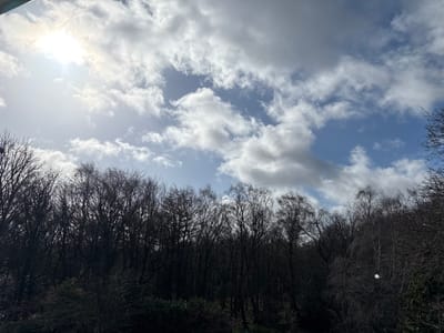The scene depicts a natural wooded area in Kirklees, United Kingdom, under a partly cloudy sky during daytime. The upper two-thirds of the image show a bright blue sky with numerous white cumulus clouds. The sun is visible in the upper-left quadrant, producing a strong glare. The lower third of the image is dominated by a dense, dark mass of predominantly leafless deciduous trees, suggesting late autumn or winter conditions. Some evergreen foliage is visible at the very bottom of the frame, beneath the taller trees. A small, circular, bright white light artifact or reflection is present within the tree line in the lower-right area. No individuals, animals, or discernible man-made structures are present within the frame.