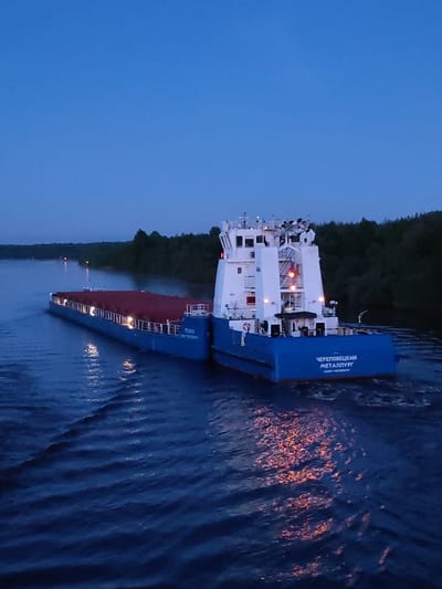 A blue and white river barge travels down a river at dusk. The barge's name, "ЧЕРЕПОВЕЦКИЙ МЕТАЛЛУРГ," is written on the stern, along with "САНКТ-ПЕТЕРБУРГ." The barge has a red cargo area, and its lights reflect on the river's surface. Trees line the riverbanks. The image was taken in Annensky Most, Russia.