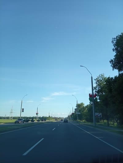 The image captures a view from inside a vehicle traveling on a multi-lane road, likely a highway, on a clear day with a pale blue sky. In the foreground, the asphalt road is marked with white lane lines. The road is flanked by grassy verges and trees on the right. On the left side of the road, there are fields and a large power pylon.
Several streetlights are visible along the road, some decorated with red and green flags. There are also decorative planters with bird-like structures along the roadside. A sign with Cyrillic text, possibly indicating a town or region, is visible on the right side. Traffic is light, with a few cars visible on the road in the distance. The environment suggests a semi-urban or suburban setting. The location in the city of Буйніцкі сельскі Савет, Belarus is indicated by the presence of a sign with Belarusian text.