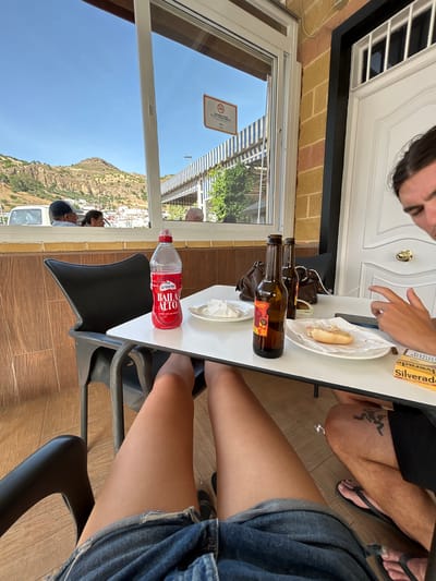 The image captures a scene at a cafe in Pizarra, Spain, likely on a sunny day. In the foreground, a person's legs in denim shorts are visible, seated at a white table. On the table are two brown beer bottles, a bottle of red liquid labeled "Baila Alto" which appears to be mineral water, a plate with a piece of food that looks like bread with a topping, and a pack of cigarettes labeled "Silverado". Another person's legs are visible on the right side of the frame, wearing flip-flops and showing a tattoo on their calf. In the background, viewed through a window, is a landscape with mountains and buildings, characteristic of Pizarra's surroundings. A striped awning is visible outside. The cafe's interior features brick walls and a white door with a gold handle. A "No Smoking" sign is affixed to the window. The overall atmosphere suggests a casual outdoor dining or drinking experience.