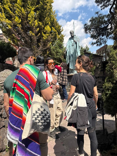 The image captures a gathering of several people in the city of San Mateo Ozolco, Mexico. They are situated near a stone statue depicting a robed figure holding a staff, possibly a religious or historical figure. The group appears to be resting or socializing on a stone pathway. One person is wearing a brightly colored, striped shawl, while another is draped with a light-colored cloth featuring a pattern of birds and fish. A woman stands wearing a black shirt and leggings with a jacket, looking towards the gathering. The backdrop includes trees and a partly cloudy sky. The environment suggests an outdoor setting, potentially a park or historical site.