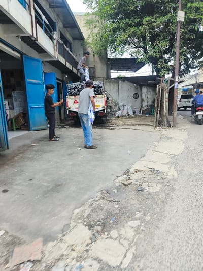 Three men are unloading materials from a white truck parked beside a building with open blue doors. One man stands on the truck bed, handing packages to a second man, while the third man stands beside the open doors, presumably taking the packages. A street runs along the side with a motorcycle and a white car visible in the background. The area appears to be a commercial or industrial zone with buildings and storage spaces nearby.