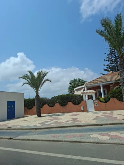 In La Manga Del Mar Menor, Spain, on a clear, sunny day with scattered clouds, a residential property is visible. The scene is viewed from a street, looking towards a house with a terracotta-tiled roof and orange-painted walls. A white gate leads to the property, which is bordered by a low, undulating orange wall topped with dense green bushes. A single palm tree stands in front of the wall, with another palm tree and a coniferous tree visible behind the house. To the left of the house, a simple white structure with a bright blue door is present. The foreground shows a road with distinct paving patterns and sidewalks. There are no people or animals visible in the image.