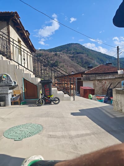 The image shows an outdoor scene in what appears to be a residential area. In the background, a forested mountain rises against a blue sky with scattered clouds. Closer in the frame, there is a two-story building with a tile roof and a metal railing along a staircase. Adjacent to the staircase is a small child's balance bike and a black garbage container. Next to the building a small yard appears to have various items. A manhole cover is visible in the foreground. The location is Grohotno, Bulgaria.