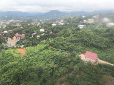 This is an aerial, eye-level, wide shot of a lush, green, hilly landscape with scattered buildings and homes, taken on an overcast day. The scene is likely in or near the area of ქვედა სამება, Georgia. In the foreground, dense green trees and foliage cover the slopes. A red-roofed house is visible on the right side of the frame, situated on a hillside with a dirt road leading to it. Further into the scene, on the left, a stone building complex with orange-tiled roofs is present, resembling a monastery or historical structure. In the background, more homes and buildings are scattered across the hills, extending to the horizon where mist-covered mountains loom under a cloudy sky. The overall impression is one of a rural or semi-rural settlement nestled within a verdant, mountainous terrain.