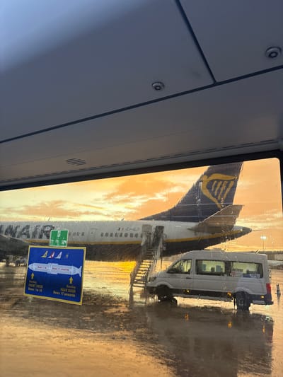 The image, taken from inside an aircraft, shows a Ryanair plane visible through a rain-streaked window. The tail of the Ryanair plane features the airline's distinctive blue and yellow logo. A white passenger transport vehicle is parked alongside the plane. A sticker on the window provides boarding instructions with directional arrows, indicating "FRONT DOOR Rows 1 to 16" and "REAR DOOR Rows 17 onwards". This indicates a Ryanair flight boarding process. The warm, golden-hued sky suggests either sunrise or sunset. The setting is likely Elx / Elche, Spain, as Ryanair operates flights from Alicante–Elche Miguel Hernández Airport (ALC) that serves the city.