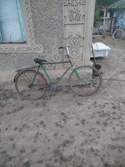In Leușeni, Moldova, an old, green bicycle is parked on a dirt path next to a textured concrete wall with decorative carvings. The bicycle has a worn black seat, a rusty rear fender, and spoked wheels with reddish-brown rims. The front wheel is turned towards the right. The wall features intricate, symmetrical patterns carved into the concrete. To the right of the bicycle, a white, tarp-covered object rests on the ground, and further back, another bicycle is visible in the distance. The ground is uneven and covered with dirt, small stones, and sparse patches of grass. The overall scene suggests an outdoor setting, possibly a yard or alleyway in a rural or semi-rural area.
