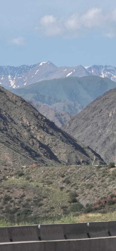 The image depicts a mountainous landscape in the vicinity of Sasyk-Bulak, Kyrgyzstan. In the foreground, a concrete barrier with white triangular markers lines the bottom of the frame. Beyond the barrier, a sloped terrain covered in dry grass and sparse vegetation rises towards the middle ground. Two steep, rocky mountainsides flank a valley, with a visible road winding through it. A power pole or transmission tower is visible on the left mountainside. In the background, successive layers of mountains fade into the distance, with the furthest peaks showing patches of snow. The sky above is a clear blue with a few scattered white clouds. The scene is characterized by its rugged terrain, arid vegetation, and distant snow-capped mountains, indicative of a high-altitude, possibly semi-arid environment. There are no people or animals visible in the image.