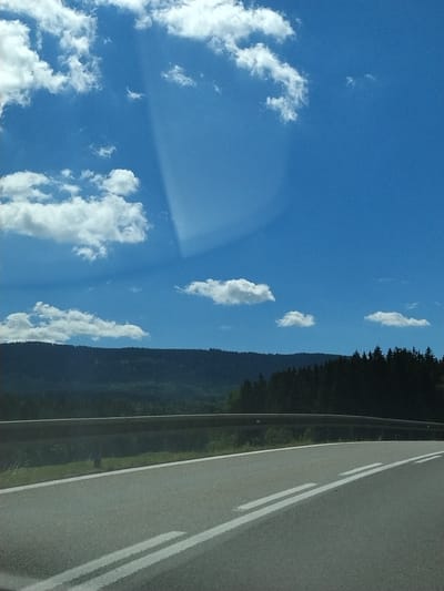 The image depicts a road scene with a wide blue sky and scattered white clouds. A dark, tree-covered hill or mountain range forms the horizon in the background. The road itself is paved and has white lane markings. A metal guardrail runs along the side of the road. The overall impression is a scenic view from a moving vehicle, likely on a highway or a scenic route. There are no people or specific landmarks of Vimperk, Czechia visible in the image.