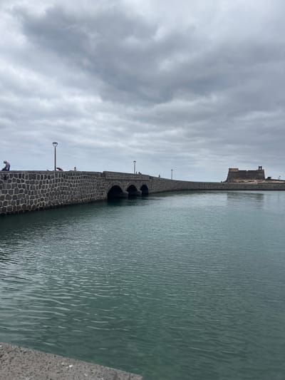 The image shows the Castillo de San Gabriel in Arrecife, Spain. It is located on an islet connected to the city by the Puente de las Bolas, a stone bridge with multiple arches. The water surrounding the islet is calm. The sky is cloudy.