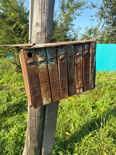 A rusted, multi-compartment mailbox unit is affixed to a wooden pole in an outdoor setting. The mailbox, likely constructed of metal, exhibits significant rust and chipping of its blue paint, with the individual compartments clearly visible. Each compartment appears to have a circular lock mechanism. Some of the compartments have faint markings or lettering on their fronts, possibly names or numbers. The mailbox unit is attached to a thick, weathered wooden post that also has a concrete support. The background consists of lush green grass and foliage, with trees visible further back. A bright turquoise corrugated metal fence runs along the right side of the scene. The sky is clear and blue. The image appears to be taken in the village of Bolshiye Shady, Russia, given the visual cues of rural or semi-rural architecture and natural surroundings. There are no people or animals visible in the frame. The lighting suggests it is daytime, with sunlight casting shadows.
