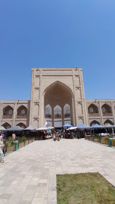 The image depicts the exterior of a large, ancient brick building with an arched entryway, likely a historical site or marketplace in Denov, Uzbekistan. The architecture features several smaller arched windows and decorative brickwork. In front of the building, a paved plaza is set up with numerous market stalls covered by various colored awnings, where vendors appear to be selling goods. Several people are visible browsing the stalls and walking in the plaza. A person is riding a small, motorized scooter-like device through the market. The sky is a clear, bright blue, indicating a sunny day. The ground is paved with large, light-colored rectangular stones, and there is a patch of green grass to the right of the plaza.