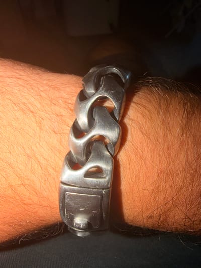 A close-up, eye-level shot shows a thick, silver-colored metal bracelet with large, interlocking links worn on a person's wrist. The bracelet is positioned horizontally across the frame, with the clasp visible on the left side. The wrist is covered in fair skin with visible hair. The background is dark and out of focus, suggesting an indoor setting. There is no indication of the location being Castillo Bajo-conejito, Spain.