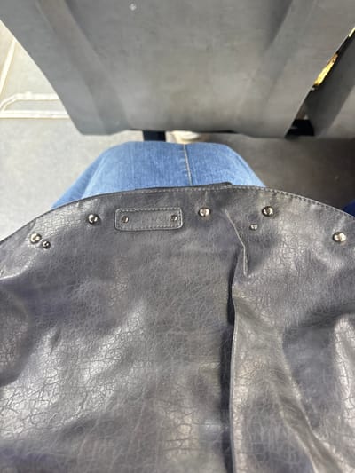 A top-down view captures a static scene within a public transport vehicle in José C. Paz, Argentina. In the immediate foreground, a dark grey faux leather bag rests on the lap of an individual, whose blue denim-clad legs are visible beneath it. The bag features a textured, slightly worn surface adorned with multiple silver-toned studs along its curved upper edge. A rectangular label, embossed with the word 'SPINOLO,' is affixed near the bag's top center, secured by two small rivets. Behind the individual, two unoccupied, worn grey seats, consistent with public transportation seating, are partially visible. A metal railing is present on the far left, and a sliver of light-colored background or exterior is visible through a gap in the seating on the right. No discernible actions or interactions are depicted.