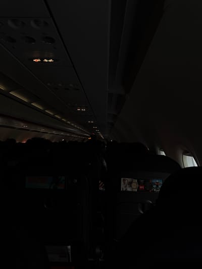 The image shows the interior of an airplane cabin. The aisle is visible, with rows of seats on either side. Overhead compartments line the upper part of the cabin. There are lights and vents visible along the cabin wall. In the foreground, the back of a seat is visible with an in-flight entertainment screen. The location is not in Peschiera Borromeo, Italy.