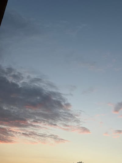 The image shows a sky with clouds during either sunrise or sunset. The sky transitions from a pale yellow at the bottom to a light blue at the top. Wispy clouds are visible throughout the sky, with some areas showing darker, more defined clouds tinged with pink and orange hues. The top left corner of the image shows a dark, vertical object, possibly a building or structure. There are no people or specific landmarks visible in the image that would indicate its location in Marki, Poland.