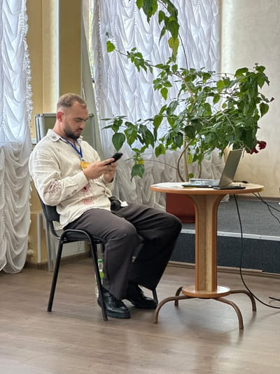 In this media file, a man is seated in a black chair, leaning back and looking down at his phone, which he holds in both hands. He is wearing a light-colored shirt with dark embroidered designs and dark trousers. A blue lanyard with a yellow badge is around his neck. He has a beard and short hair. The scene appears to be indoors, with a wooden floor. To his left, a large potted plant with green leaves and a few red flowers is prominent. Behind him, a round wooden table with a light-colored top and dark metal legs is visible, with a laptop on it. The background shows white curtains and a hint of a window. The overall lighting suggests daytime. The image is oriented vertically, with the man appearing in the lower portion of the frame.
