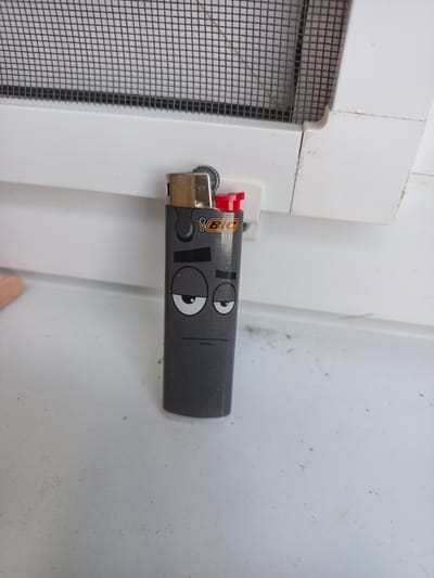 A Bic lighter with a cartoonish face printed on it sits on a white surface, possibly a windowsill, next to a window screen. The lighter is gray with a gold top and a red ignition button. The face on the lighter has two large, droopy eyes and a thin, horizontal line for a mouth. The window screen is black and has a grid pattern. The background is out of focus but appears to be a white wall.