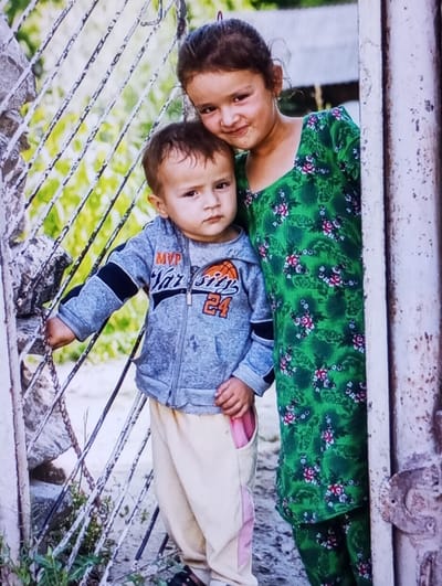 The image displays two children, a girl and a boy, outdoors in Yangirabat, Uzbekistan. The older child, a girl estimated to be 5-7 years old, with dark hair, wears a green dress adorned with a repetitive pattern of red, pink, and white flowers and green leaves. She stands on the right, her head resting on the younger boy's head, and her left arm around his shoulder. The younger child, a boy estimated to be 1-2 years old, wears a grey zippered hoodie with black sleeves, featuring "MVP," "VARSITY," an orange basketball graphic, and the number "24." He also wears light beige sweatpants with pink side panels. His left hand is grasping a distressed, light-colored metal gate with horizontal and diagonal bars, located to his left. To the right of the children, a weathered, vertical post or door frame is visible. The background features blurred green foliage and an unpaved ground surface, illuminated by natural daylight.