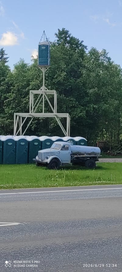 In Võru linn, Estonia, a row of green portable toilets is lined up on a grassy area. Behind them, a light blue vintage truck with a large tank on its back is parked. Further behind the truck and toilets, a tall, white metal structure supports a single green portable toilet at its apex, surrounded by lush green trees under a clear blue sky with scattered clouds. A paved road is visible in the foreground.