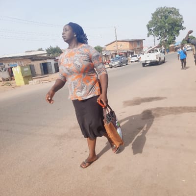 The image depicts a woman walking down a street in Kufang, Nigeria (latitude: 9.863, longitude: 8.867). She is wearing a long-sleeved patterned shirt and a black skirt, and carrying a large brown bag. She is walking in the direction of the camera, towards the right side of the frame. There are three parked cars and a white pickup truck in the background, and a few other buildings. The road is paved and has a slight incline. The environment is sunny and dry. There are other people in the background, walking in the opposite direction, but they are not in focus. The woman appears to be alone, and the scene is relatively quiet.