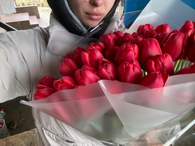 An individual is depicted holding a large bouquet of approximately 50-70 vibrant red tulips outdoors in Trudobelikovskiy, Russia. The person's lower face, including the mouth and nose, is visible. They are wearing a light-colored, possibly beige or light grey, puffer jacket and a dark hooded garment. The tulips are tightly arranged and wrapped in translucent white paper and clear plastic film. In the background, stone or concrete steps ascend to the left. The ground surface is paved with hexagonal tiles. A red-capped plastic bottle and a small brown basket or container are visible on the ground to the lower left, adjacent to a wooden surface. A blue structure is partially visible in the upper right background.